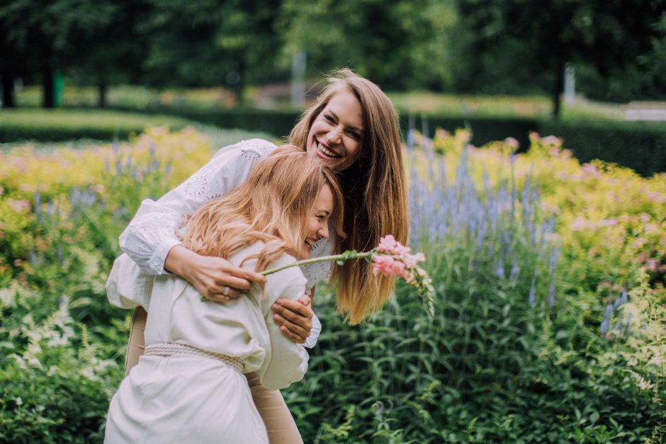 A woman and a young girl are outdoors in a lush garden with green foliage and a variety of blooming flowers in pastel shades. The woman has long, light brown hair and is smiling joyfully as she embrac